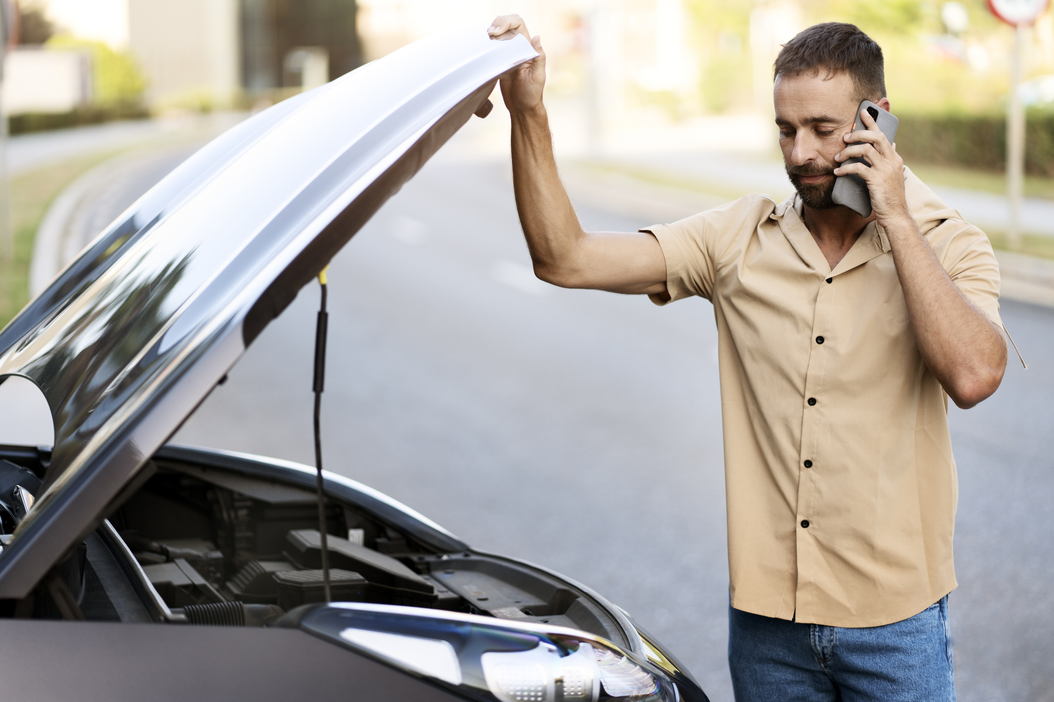 Sad driver calling car service, opening hood, having engine problem standing near broken car on the road. Car breakdown concept